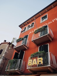 A red building with multiple balconies adorned with green plants. Signs for 'HOTEL' and 'BAR' are prominently displayed on the facade. The building features dark green shutters and architectural details reminiscent of European style.