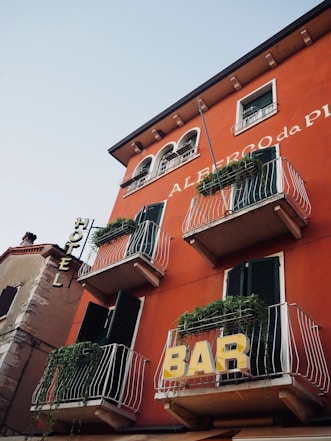 A red building with multiple balconies adorned with green plants. Signs for 'HOTEL' and 'BAR' are prominently displayed on the facade. The building features dark green shutters and architectural details reminiscent of European style.