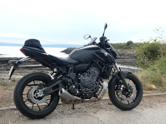 A sleek black motorcycle is parked on a coastal road with rocky terrain and sparse vegetation surrounding it. The sea is visible in the background, under a cloudy sky.