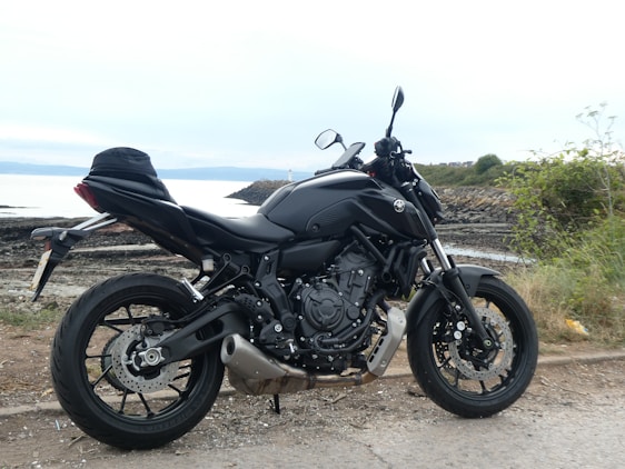 A sleek black motorcycle is parked on a coastal road with rocky terrain and sparse vegetation surrounding it. The sea is visible in the background, under a cloudy sky.