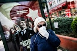 A person wearing a white beanie, sunglasses, and a face mask stands in front of a public advertisement featuring historical figures in formal clothing. The advertisement includes a large Turkish flag and contains text in Turkish. The scene appears to be set in an urban outdoor environment, with buildings and greenery visible in the background.
