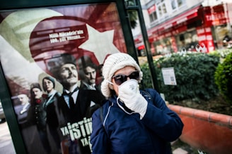 A person wearing a white beanie, sunglasses, and a face mask stands in front of a public advertisement featuring historical figures in formal clothing. The advertisement includes a large Turkish flag and contains text in Turkish. The scene appears to be set in an urban outdoor environment, with buildings and greenery visible in the background.