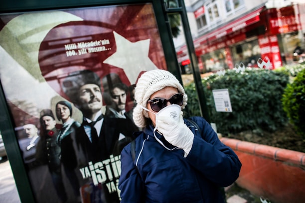 A person wearing a white beanie, sunglasses, and a face mask stands in front of a public advertisement featuring historical figures in formal clothing. The advertisement includes a large Turkish flag and contains text in Turkish. The scene appears to be set in an urban outdoor environment, with buildings and greenery visible in the background.