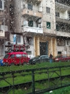 Raindrops cover a window, creating a pattern on the glass. Behind the window, a residential street scene features a red van parked alongside other cars in front of an apartment building. The building displays tones of beige and gray, with balconies and windows visible. Green grass is in the foreground.