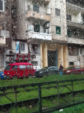 Raindrops cover a window, creating a pattern on the glass. Behind the window, a residential street scene features a red van parked alongside other cars in front of an apartment building. The building displays tones of beige and gray, with balconies and windows visible. Green grass is in the foreground.