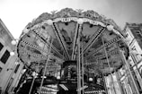 A classic carousel with intricate designs and paintings on the underside of the canopy. The structure is adorned with numerous lights, and several vertical poles are visible. The background includes parts of nearby buildings, indicating an urban setting.