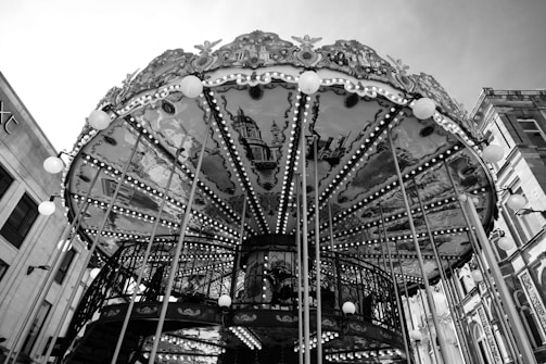 A classic carousel with intricate designs and paintings on the underside of the canopy. The structure is adorned with numerous lights, and several vertical poles are visible. The background includes parts of nearby buildings, indicating an urban setting.