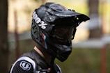 A person wearing a black full-face helmet with large, clear goggles. The helmet has branding and colorful stripes, and the person appears to be dressed in a matching sports outfit with logos. The lighting is dim, casting shadows across the person's face.