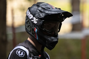 A person wearing a black full-face helmet with large, clear goggles. The helmet has branding and colorful stripes, and the person appears to be dressed in a matching sports outfit with logos. The lighting is dim, casting shadows across the person's face.