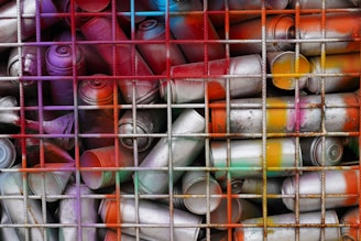 A collection of spray paint cans is packed into a grid-like wire structure. The cans are variously colored with remnants of orange, purple, yellow, red, and blue paint. Some cans appear to be metallic and worn, suggesting they may have been used multiple times.