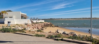 A serene coastal scene featuring a white building with a shaded patio on the left adjacent to sandy beach and rocky shoreline. The foreground shows a mix of concrete, rocks, and green vegetation. The expansive sky is clear with soft clouds, and the ocean is gently lapping against the shore. There is a lamppost and a trash bin nearby.