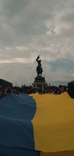 A group of people hold a large Ukrainian flag in a public square, with a prominent statue in the background against a cloudy sky.
