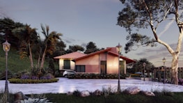 A modern house with a sloped red tile roof surrounded by lush green trees and landscaping. The house features large windows and a well-maintained garden area. A driveway leads up to the house with vintage-style street lamps lining the path.