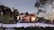 A modern house with a sloped red tile roof surrounded by lush green trees and landscaping. The house features large windows and a well-maintained garden area. A driveway leads up to the house with vintage-style street lamps lining the path.