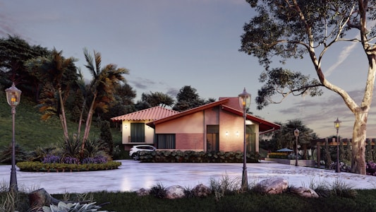 A modern house with a sloped red tile roof surrounded by lush green trees and landscaping. The house features large windows and a well-maintained garden area. A driveway leads up to the house with vintage-style street lamps lining the path.