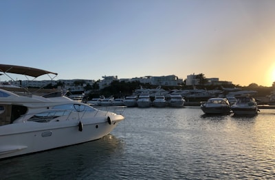 A tranquil marina at sunset with several luxury yachts parked along the water. The sky is clear with a soft gradient from blue to orange as the sun sets behind a line of modern buildings and palm trees in the distance.