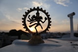 A brass figurine of the Hindu deity Shiva Nataraja is silhouetted against a setting sun in the background. The statue is intricately designed, featuring Shiva in a dance pose surrounded by a circular frame. The sky displays soft, pastel-colored clouds contrasting with the dark outline of the urban landscape below.