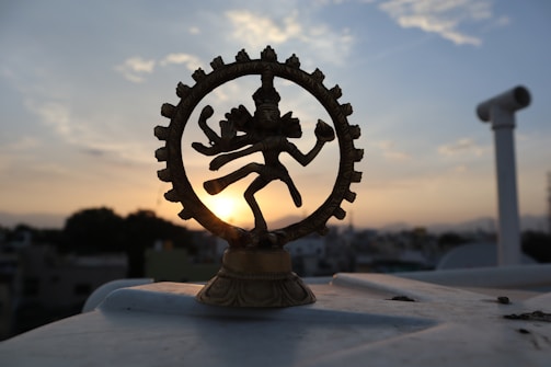 A brass figurine of the Hindu deity Shiva Nataraja is silhouetted against a setting sun in the background. The statue is intricately designed, featuring Shiva in a dance pose surrounded by a circular frame. The sky displays soft, pastel-colored clouds contrasting with the dark outline of the urban landscape below.