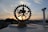 A brass figurine of the Hindu deity Shiva Nataraja is silhouetted against a setting sun in the background. The statue is intricately designed, featuring Shiva in a dance pose surrounded by a circular frame. The sky displays soft, pastel-colored clouds contrasting with the dark outline of the urban landscape below.