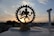 A brass figurine of the Hindu deity Shiva Nataraja is silhouetted against a setting sun in the background. The statue is intricately designed, featuring Shiva in a dance pose surrounded by a circular frame. The sky displays soft, pastel-colored clouds contrasting with the dark outline of the urban landscape below.