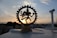 A brass figurine of the Hindu deity Shiva Nataraja is silhouetted against a setting sun in the background. The statue is intricately designed, featuring Shiva in a dance pose surrounded by a circular frame. The sky displays soft, pastel-colored clouds contrasting with the dark outline of the urban landscape below.
