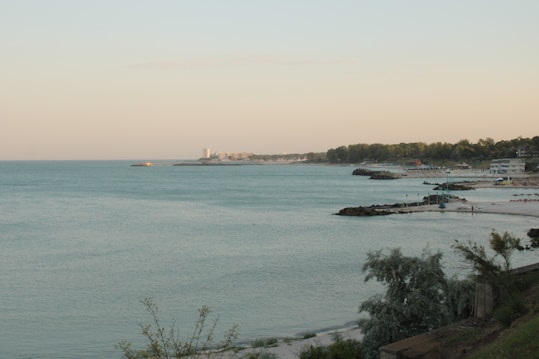 A serene coastal landscape with calm, blue waters stretching out towards the horizon. The shoreline curves gently, lined with small rocky jetties and a light sandy beach. Trees and sparse vegetation can be seen along the edge, and there are a few distant buildings along the coast, including a tall structure and lower buildings. The sky is a soft gradient of pastel colors, suggesting an early morning or late afternoon setting.