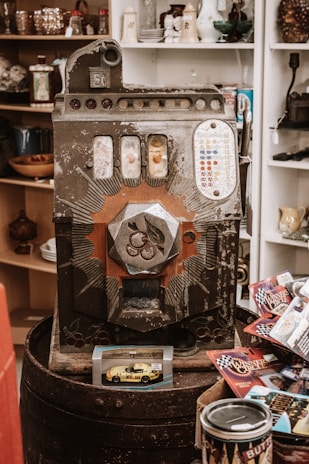 An old vintage slot machine with ornate designs and various small items surrounding it, including a toy car model in packaging and several racing-themed cards. Shelves in the background contain various decorative items such as glasses and porcelain.