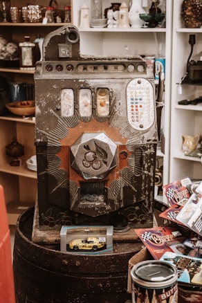 An old vintage slot machine with ornate designs and various small items surrounding it, including a toy car model in packaging and several racing-themed cards. Shelves in the background contain various decorative items such as glasses and porcelain.