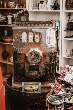 An old vintage slot machine with ornate designs and various small items surrounding it, including a toy car model in packaging and several racing-themed cards. Shelves in the background contain various decorative items such as glasses and porcelain.