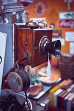 A vintage wall-mounted telephone with a wooden body, metal earpiece, and a mouthpiece. It has brass bells on top and is surrounded by various retro signs and objects like old books and a jar.