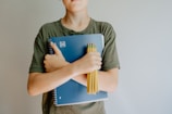 A person wearing a green shirt is holding a blue spiral notebook and a bundle of yellow pencils crossed against their chest.