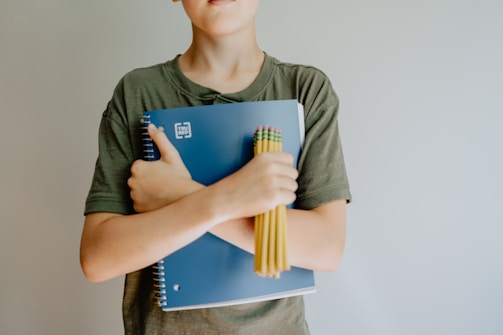 A person wearing a green shirt is holding a blue spiral notebook and a bundle of yellow pencils crossed against their chest.