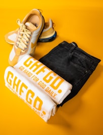 A pair of stylish yellow and white sneakers is placed alongside folded white T-shirts with bold yellow text, 'GHFGD. GO HARD FOR GOD DAILY.' A pair of dark denim jeans complements the collection, all set against a vibrant yellow background.