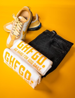 A pair of stylish yellow and white sneakers is placed alongside folded white T-shirts with bold yellow text, 'GHFGD. GO HARD FOR GOD DAILY.' A pair of dark denim jeans complements the collection, all set against a vibrant yellow background.