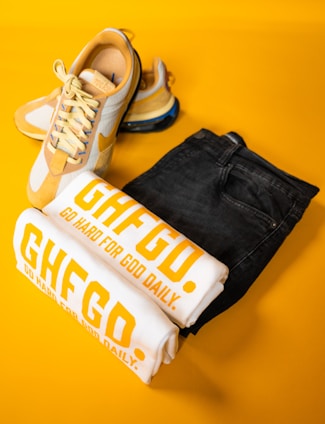 A pair of stylish yellow and white sneakers is placed alongside folded white T-shirts with bold yellow text, 'GHFGD. GO HARD FOR GOD DAILY.' A pair of dark denim jeans complements the collection, all set against a vibrant yellow background.