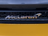 A close-up view of an automotive brand logo with a honeycomb grille pattern in the background. The logo appears metallic and reflective, positioned against a sleek, dark structure with a smooth, glossy yellow surface below.
