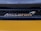 A close-up view of an automotive brand logo with a honeycomb grille pattern in the background. The logo appears metallic and reflective, positioned against a sleek, dark structure with a smooth, glossy yellow surface below.