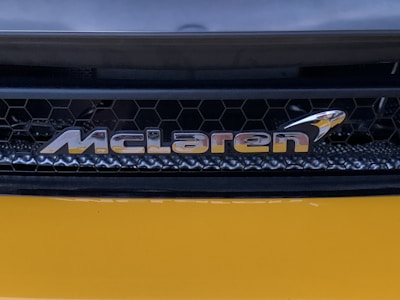 A close-up view of an automotive brand logo with a honeycomb grille pattern in the background. The logo appears metallic and reflective, positioned against a sleek, dark structure with a smooth, glossy yellow surface below.
