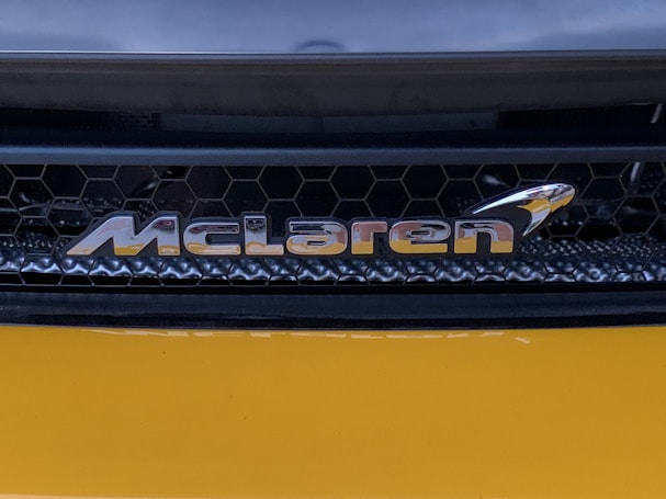 A close-up view of an automotive brand logo with a honeycomb grille pattern in the background. The logo appears metallic and reflective, positioned against a sleek, dark structure with a smooth, glossy yellow surface below.