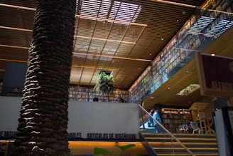 A modern bookstore interior with a large, central tree trunk reaching to the ceiling. Wooden slats above provide a warm, inviting lighting effect. Shelves filled with numerous books line the walls on multiple levels. A staircase leads to an upper level, where a plant is positioned near the railing.