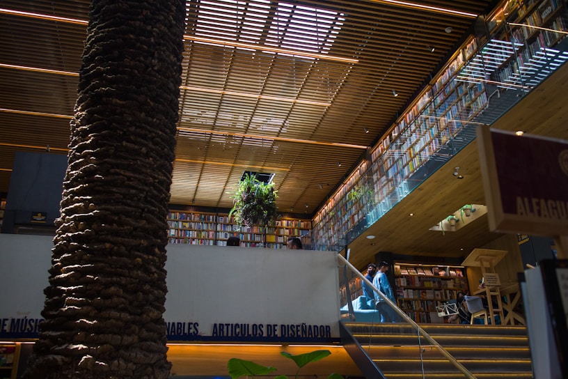 A modern bookstore interior with a large, central tree trunk reaching to the ceiling. Wooden slats above provide a warm, inviting lighting effect. Shelves filled with numerous books line the walls on multiple levels. A staircase leads to an upper level, where a plant is positioned near the railing.