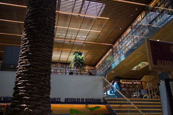 A modern bookstore interior with a large, central tree trunk reaching to the ceiling. Wooden slats above provide a warm, inviting lighting effect. Shelves filled with numerous books line the walls on multiple levels. A staircase leads to an upper level, where a plant is positioned near the railing.