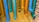 Multiple colorful pipes are attached to a yellow brick wall. The pipes are painted in vibrant shades of blue, teal, cyan, orange, and yellow, each ending with a perforated cap. The arrangement creates an artistic and playful display against the textured background.