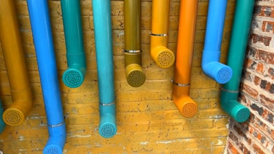 Multiple colorful pipes are attached to a yellow brick wall. The pipes are painted in vibrant shades of blue, teal, cyan, orange, and yellow, each ending with a perforated cap. The arrangement creates an artistic and playful display against the textured background.
