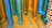 Multiple colorful pipes are attached to a yellow brick wall. The pipes are painted in vibrant shades of blue, teal, cyan, orange, and yellow, each ending with a perforated cap. The arrangement creates an artistic and playful display against the textured background.