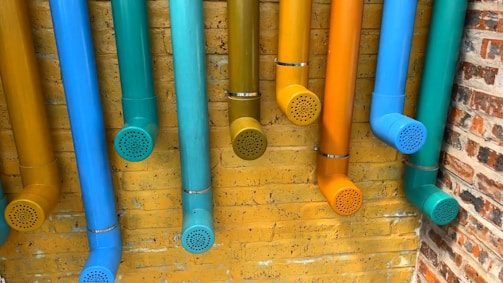 Multiple colorful pipes are attached to a yellow brick wall. The pipes are painted in vibrant shades of blue, teal, cyan, orange, and yellow, each ending with a perforated cap. The arrangement creates an artistic and playful display against the textured background.