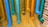 Multiple colorful pipes are attached to a yellow brick wall. The pipes are painted in vibrant shades of blue, teal, cyan, orange, and yellow, each ending with a perforated cap. The arrangement creates an artistic and playful display against the textured background.