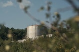 A large industrial tank stands in a natural setting surrounded by trees and foliage. The tank is cylindrical with a metallic finish, and there is a staircase leading to its top. The foreground is slightly blurred with plants and branches, while the background shows a clear blue sky with a few clouds.