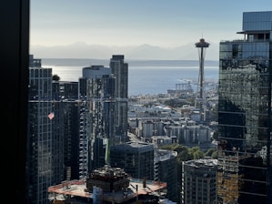 A cityscape featuring tall buildings, with a prominent tower resembling the Space Needle overlooking a body of water in the background. The scene includes construction cranes and a flag visible among the skyscrapers, while mountains are faintly visible on the horizon.