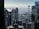 A cityscape featuring tall buildings, with a prominent tower resembling the Space Needle overlooking a body of water in the background. The scene includes construction cranes and a flag visible among the skyscrapers, while mountains are faintly visible on the horizon.